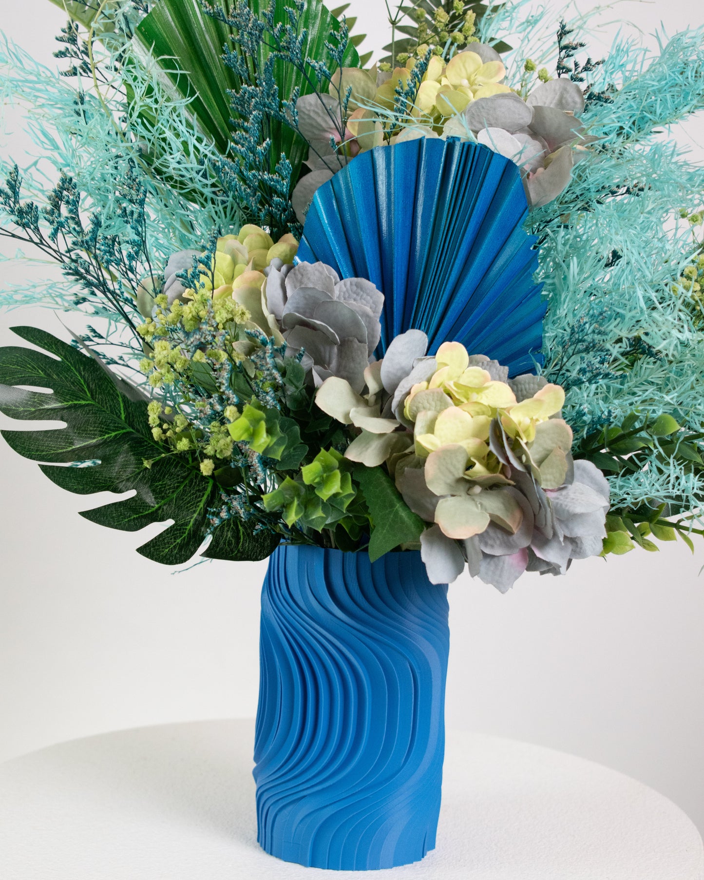 Artificial blue and green floral arrangement with tropical leaves and preserved hydrangeas in a sculptural blue vase, styled on a white pedestal against a neutral background.