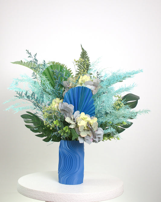 Artificial blue and green floral arrangement with tropical leaves and preserved hydrangeas in a sculptural blue vase, styled on a white pedestal against a neutral background.