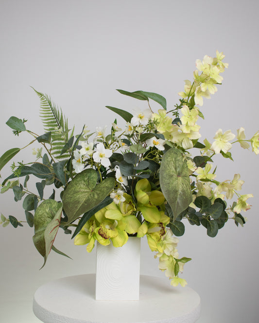 Artificial flower arrangement featuring yellow orchids, white blossoms, and lush green foliage with ferns and eucalyptus leaves, displayed in a modern white cube vase on a round white table against a neutral background.
