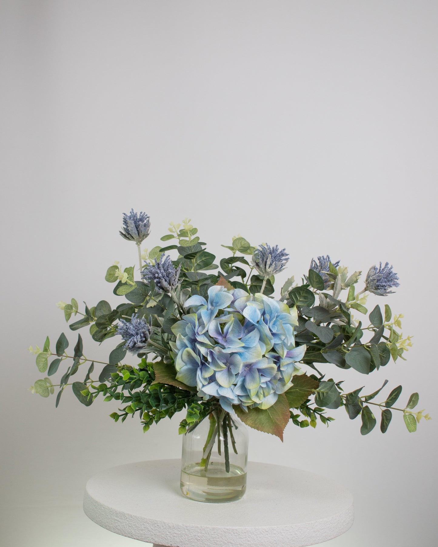 Artificial floral arrangement featuring a blue hydrangea surrounded by eucalyptus leaves and lavender-toned thistles, arranged in a clear glass vase with acrylic water, set against a neutral background.