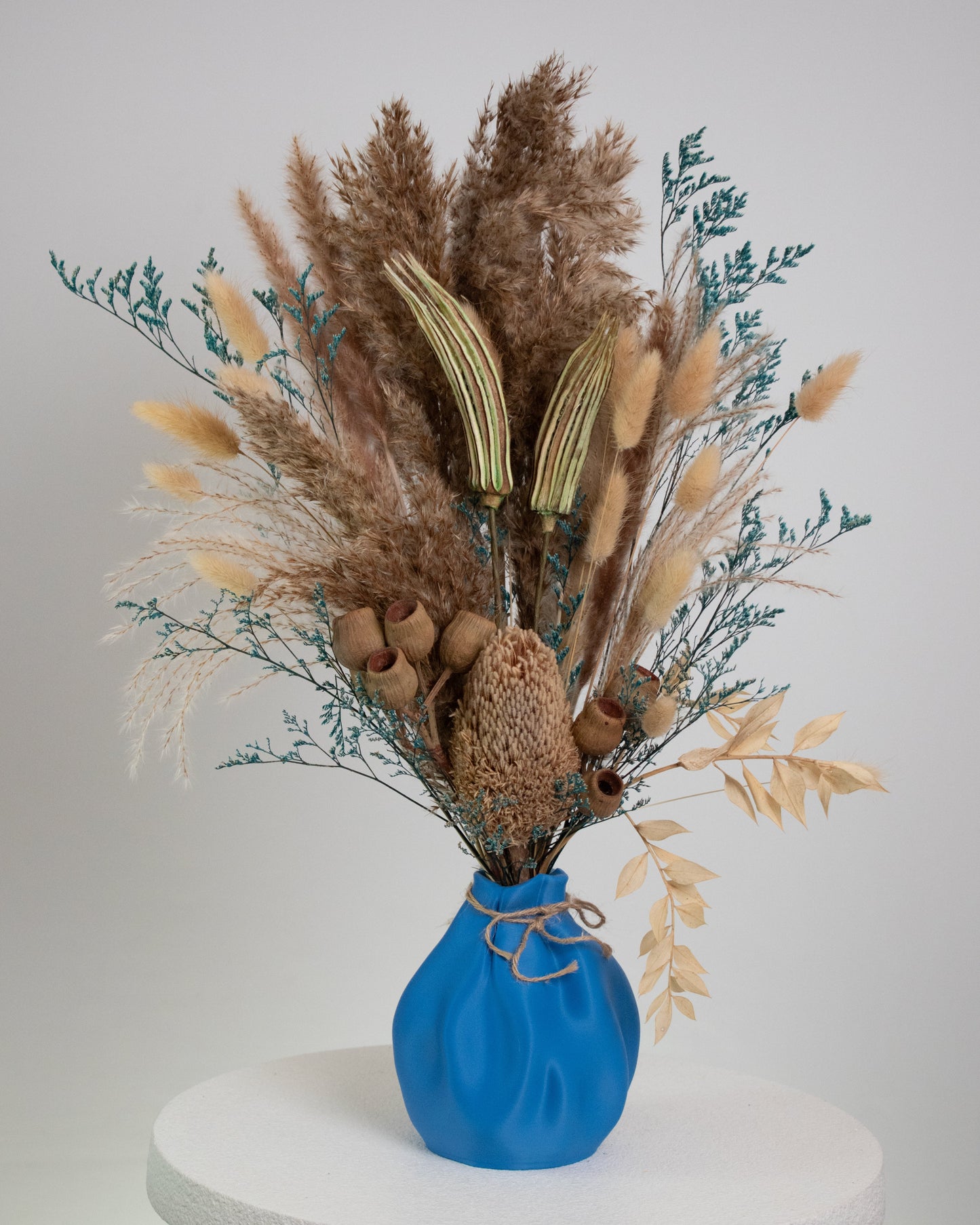 Dried floral arrangement featuring pampas grass, bunny tails, and rustic seed pods in a blue sculptural vase with jute twine, displayed on a marble pedestal.