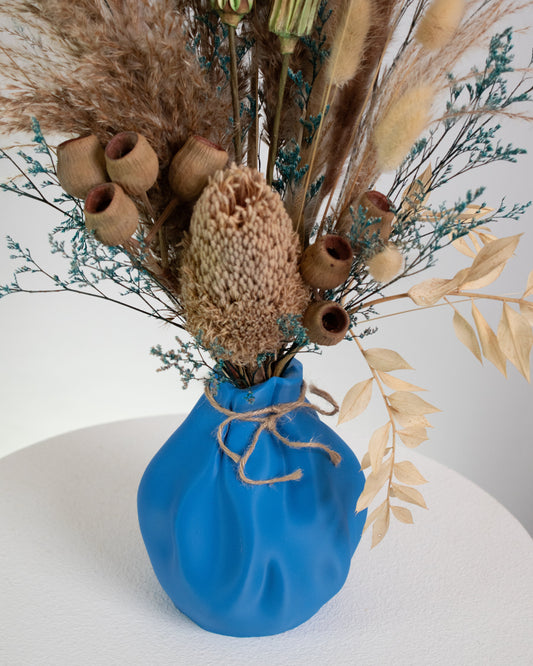 Dried floral arrangement featuring pampas grass, bunny tails, and rustic seed pods in a blue sculptural vase with jute twine, displayed on a marble pedestal.
