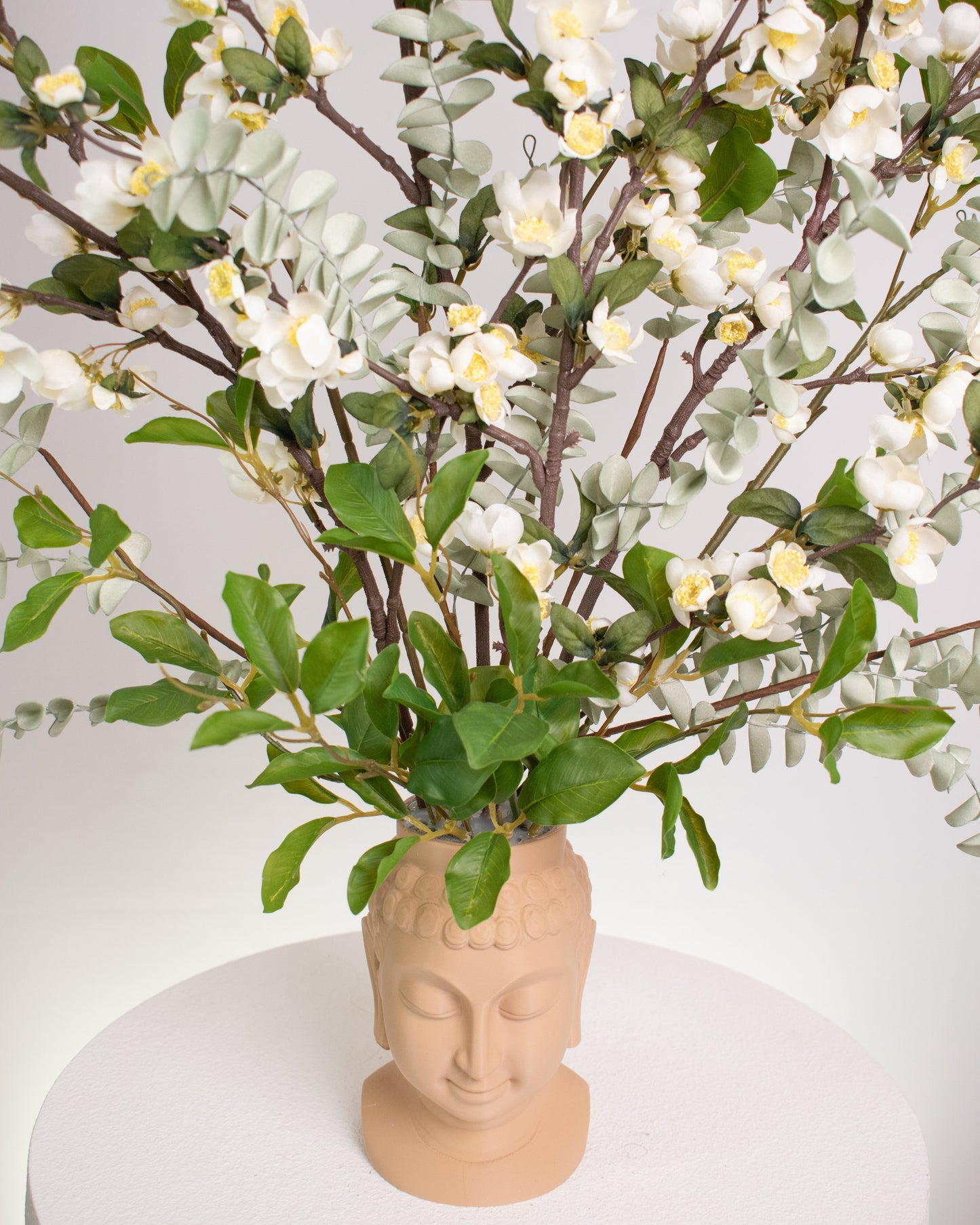Elegant artificial floral arrangement with white blossoms and green foliage displayed in a beige Buddha head vase, symbolizing calm and mindfulness in a serene design.