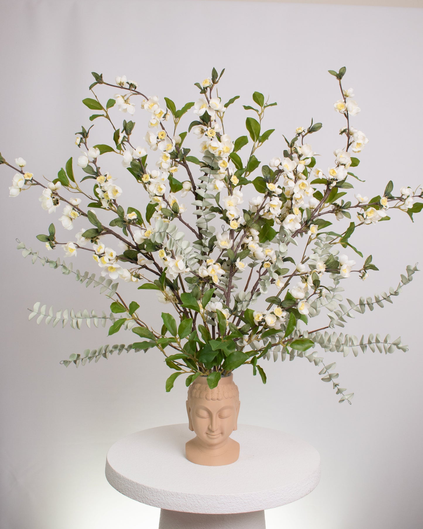 Elegant artificial floral arrangement with white blossoms and green leaves arranged in a beige Buddha head vase, symbolizing calmness and balance in modern décor.