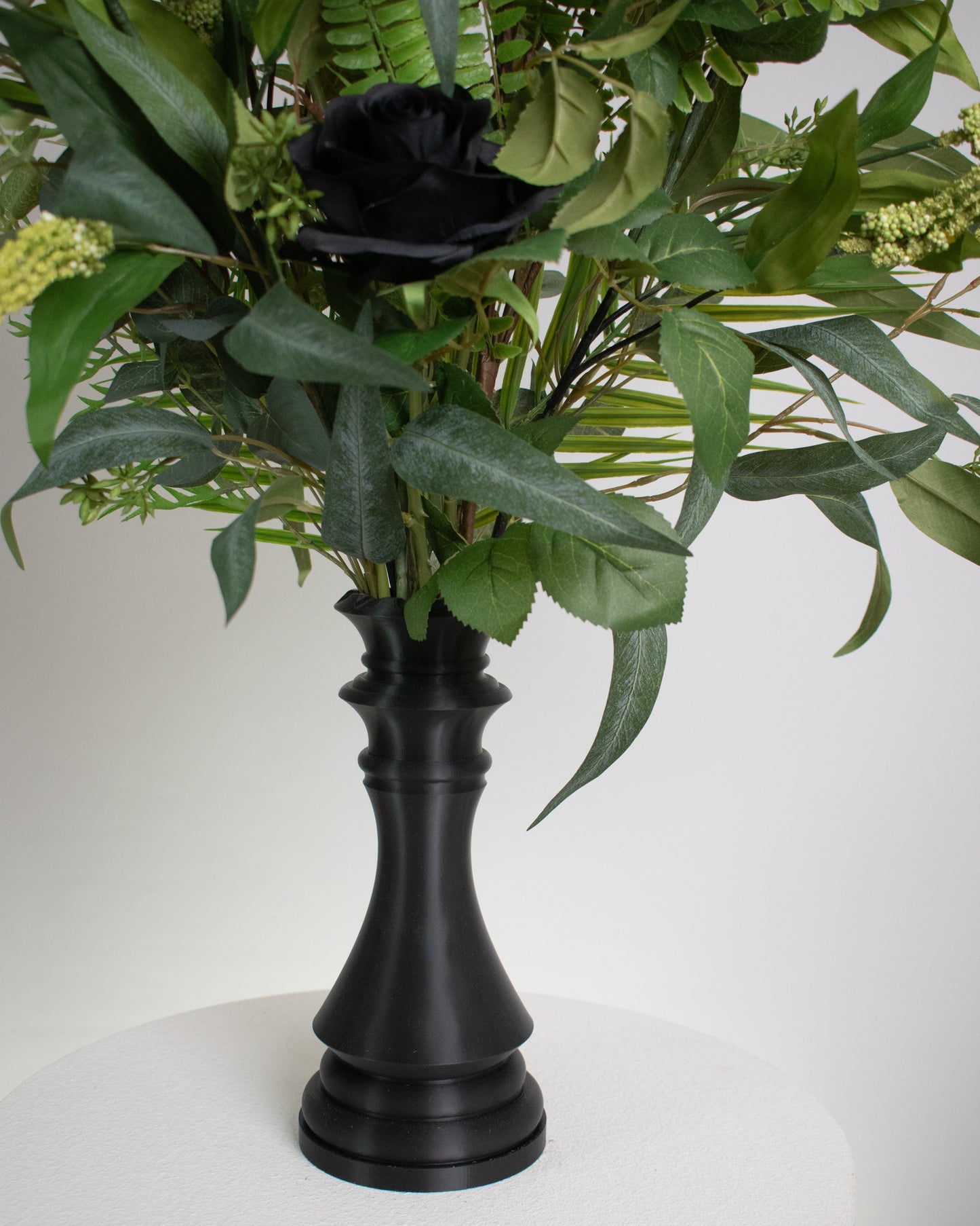 Artificial arrangement with black roses and lush green foliage displayed in a chess-queen–shaped matte-black vase, symbolizing power, sophistication, and modern elegance.