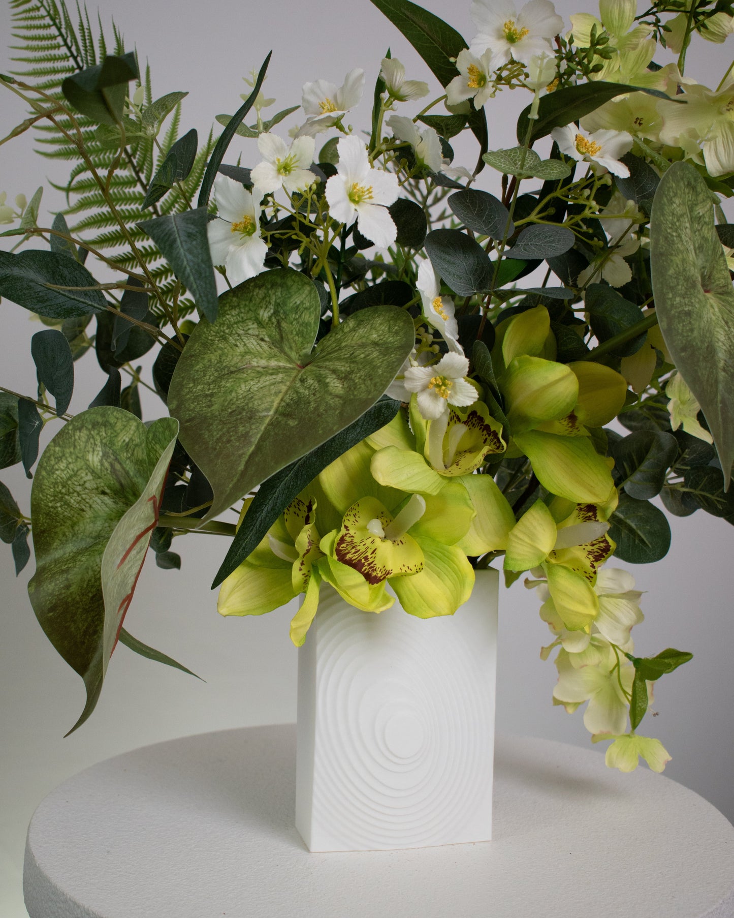 Artificial flower arrangement featuring yellow orchids, white blossoms, and lush green foliage with ferns and eucalyptus leaves, displayed in a modern white cube vase on a round white table against a neutral background.