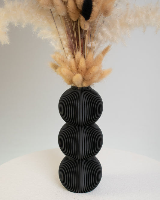A tall arrangement of dried pampas grass and bunny tails in beige and black tones, displayed in a matte black three-sphere vase on a white table.