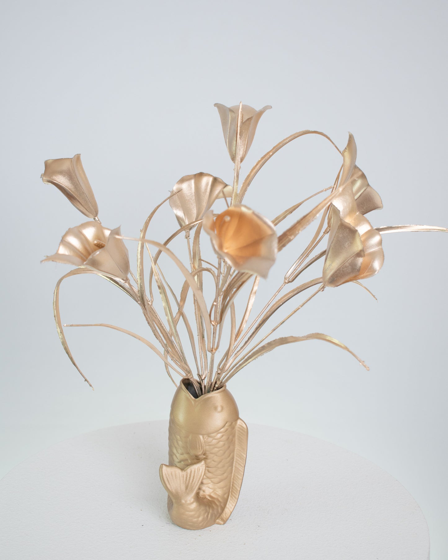 Golden metallic artificial calla lilies arranged in a gold fish-shaped vase, displayed on a white pedestal against a light neutral background.