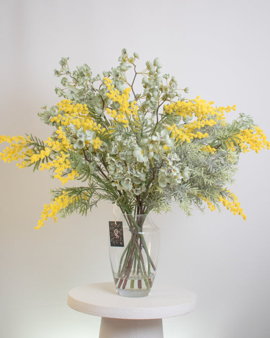 Artificial flower arrangement featuring vibrant yellow wattle blooms mixed with soft green eucalyptus leaves, displayed in a clear glass vase filled with faux stems for a natural, elegant look.