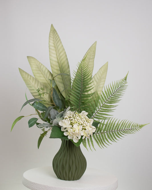 Artificial green arrangement featuring tropical leaves, ferns, eucalyptus, and ivory blooms, arranged in a textured olive-green vase on a neutral background.