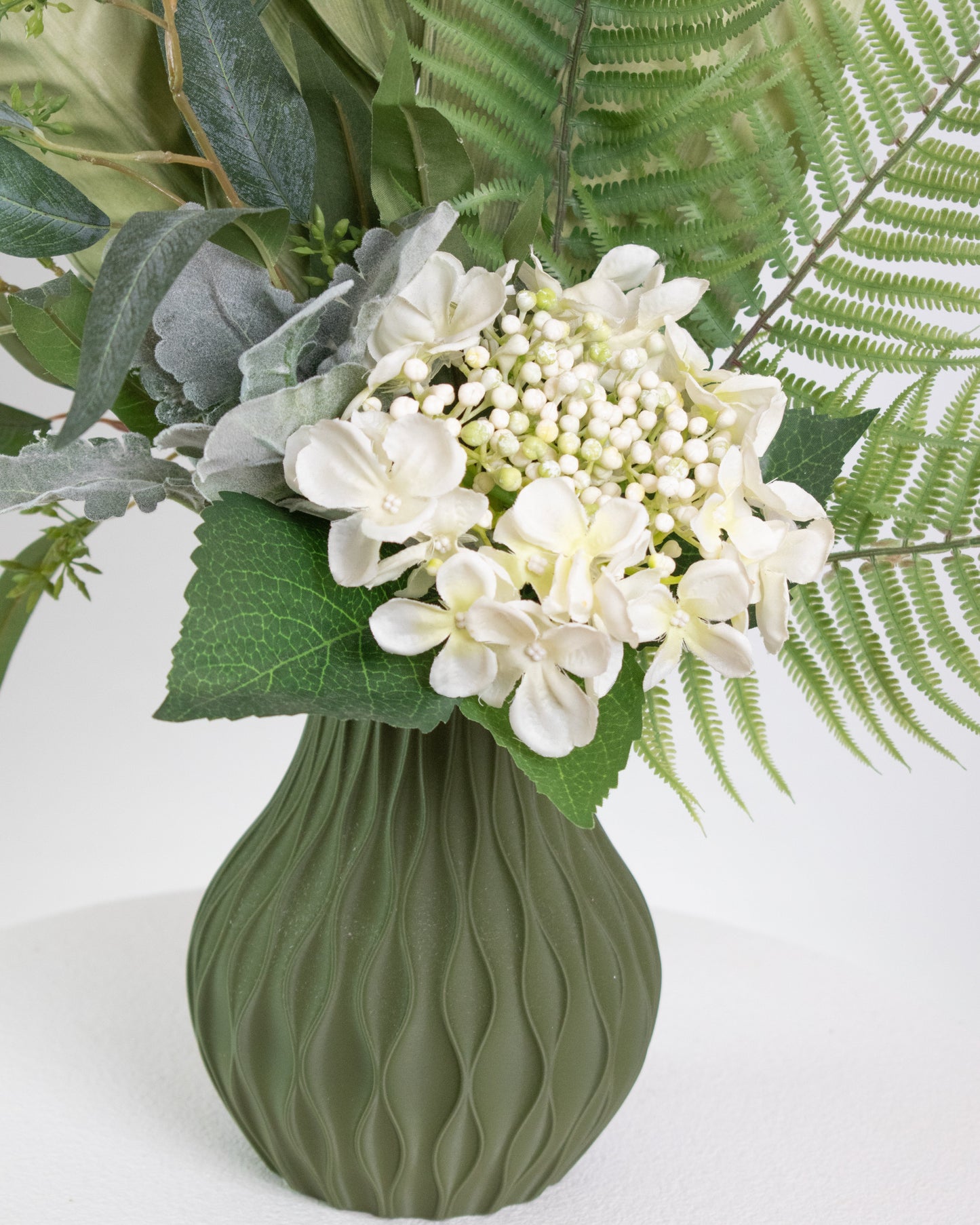 Artificial green arrangement featuring tropical leaves, ferns, eucalyptus, and ivory blooms, arranged in a textured olive-green vase on a neutral background.