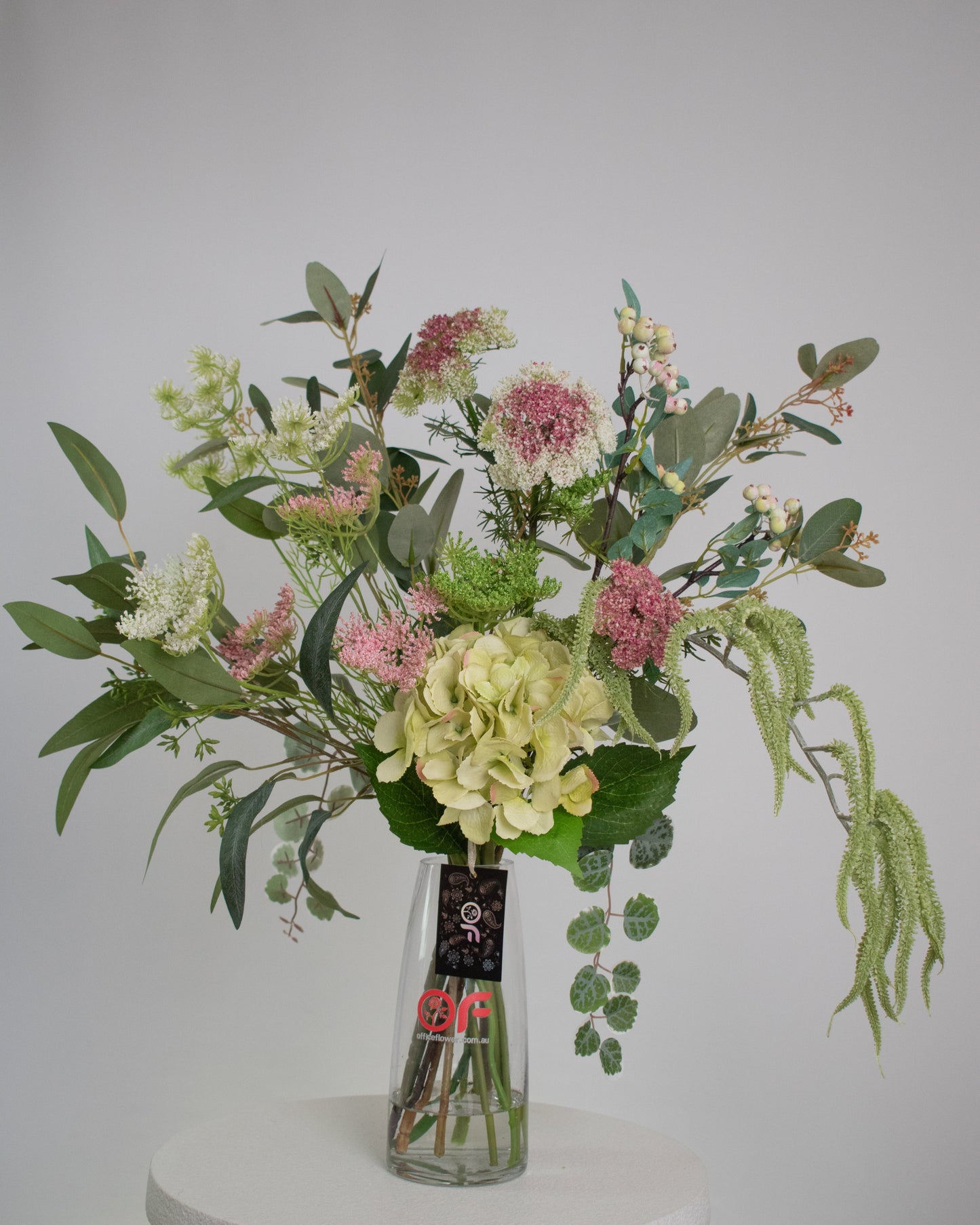 Artificial floral arrangement featuring green hydrangeas, pink wildflowers, and lush foliage in a clear glass vase with acrylic water