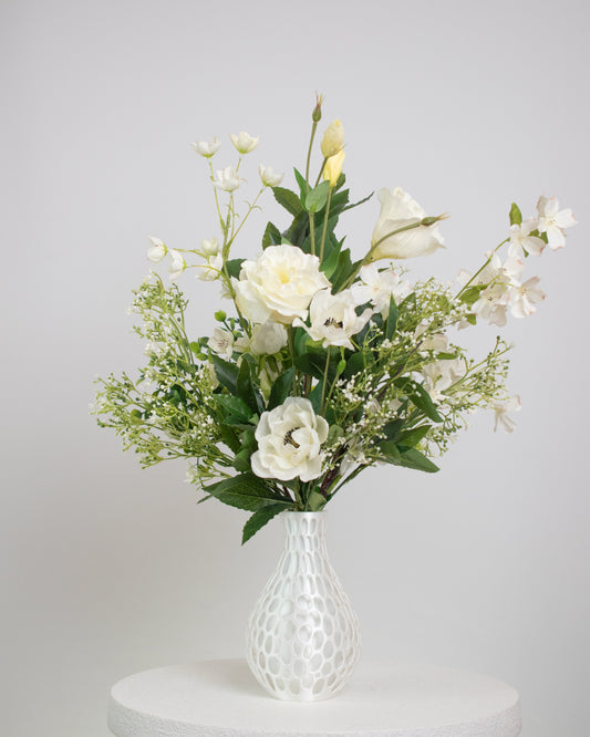 Artificial floral arrangement featuring ivory roses, white lisianthus, and blossoms with lush green foliage, styled in a white honeycomb-textured vase on a neutral background.