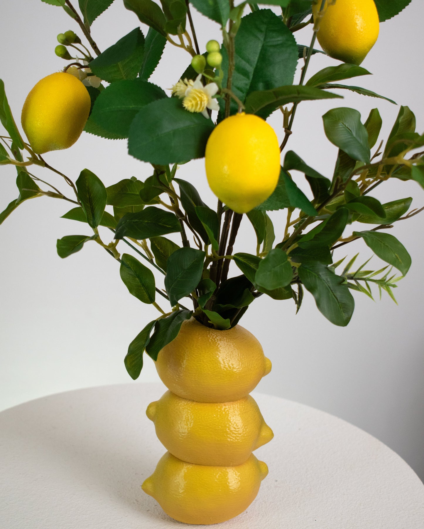 Artificial lemon arrangement featuring lifelike lemons, green leaves, and small white blossoms, displayed in a stacked lemon-shaped vase on a light background.