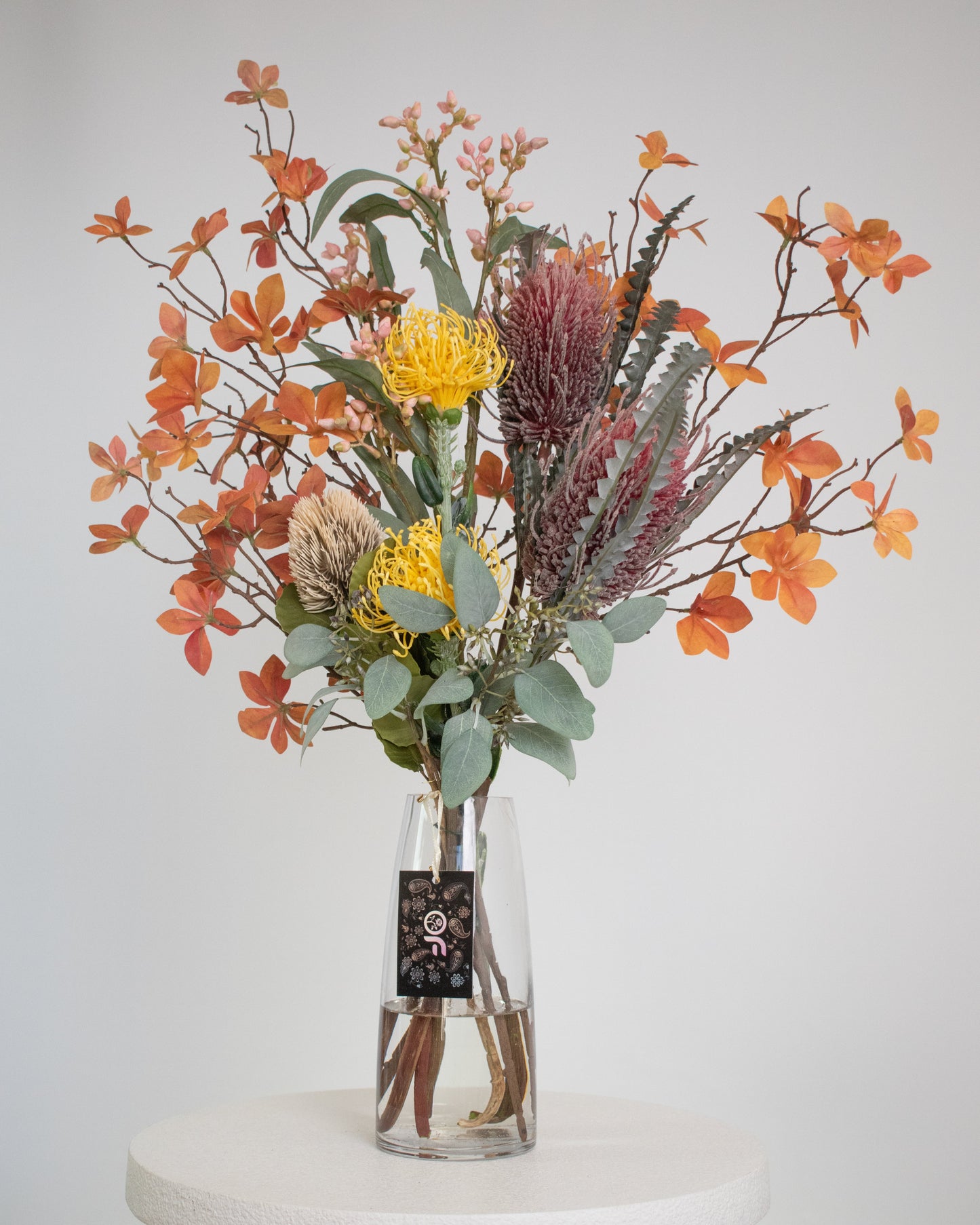 Artificial native flower arrangement featuring yellow pincushion proteas, burgundy banksia, seed pods, eucalyptus leaves, and rust-orange foliage, elegantly arranged in a clear glass vase with acrylic water.