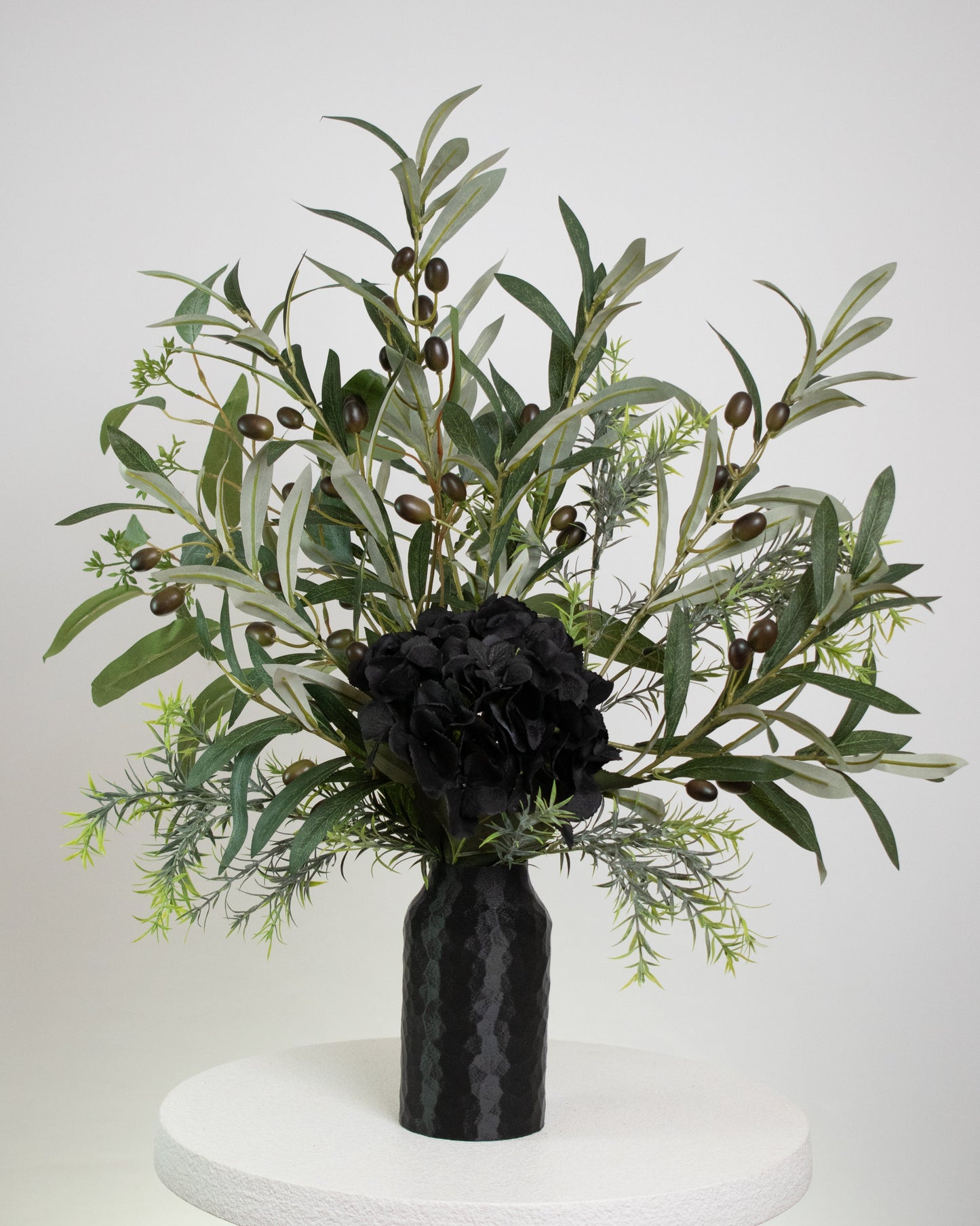Artificial floral arrangement featuring lifelike olive branches with dark berries and a black hydrangea bloom, elegantly displayed in a textured black vase on a marble-topped round table against a neutral background.