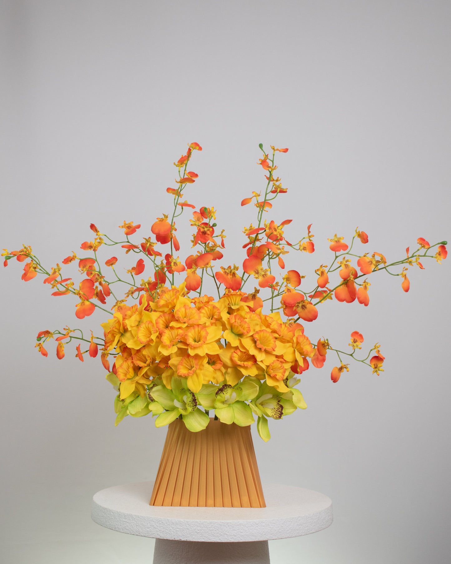 Vibrant artificial floral arrangement featuring orange orchids, yellow blossoms, and green cymbidiums in a pleated terracotta vase, styled on a white pedestal against a soft neutral background.