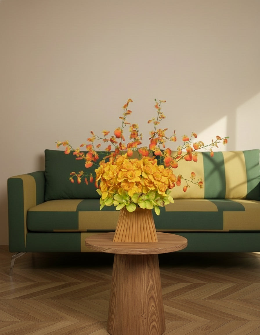 Vibrant artificial floral arrangement featuring orange orchids, yellow blossoms, and green cymbidiums in a pleated terracotta vase, styled on a white pedestal against a soft neutral background.