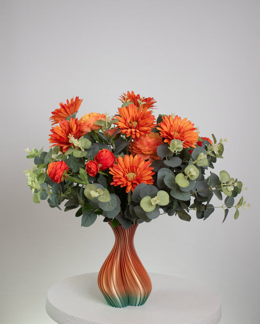 Artificial floral arrangement with orange gerberas and ranunculus surrounded by eucalyptus leaves, displayed in an ombre vase blending amber and green tones on a light background.