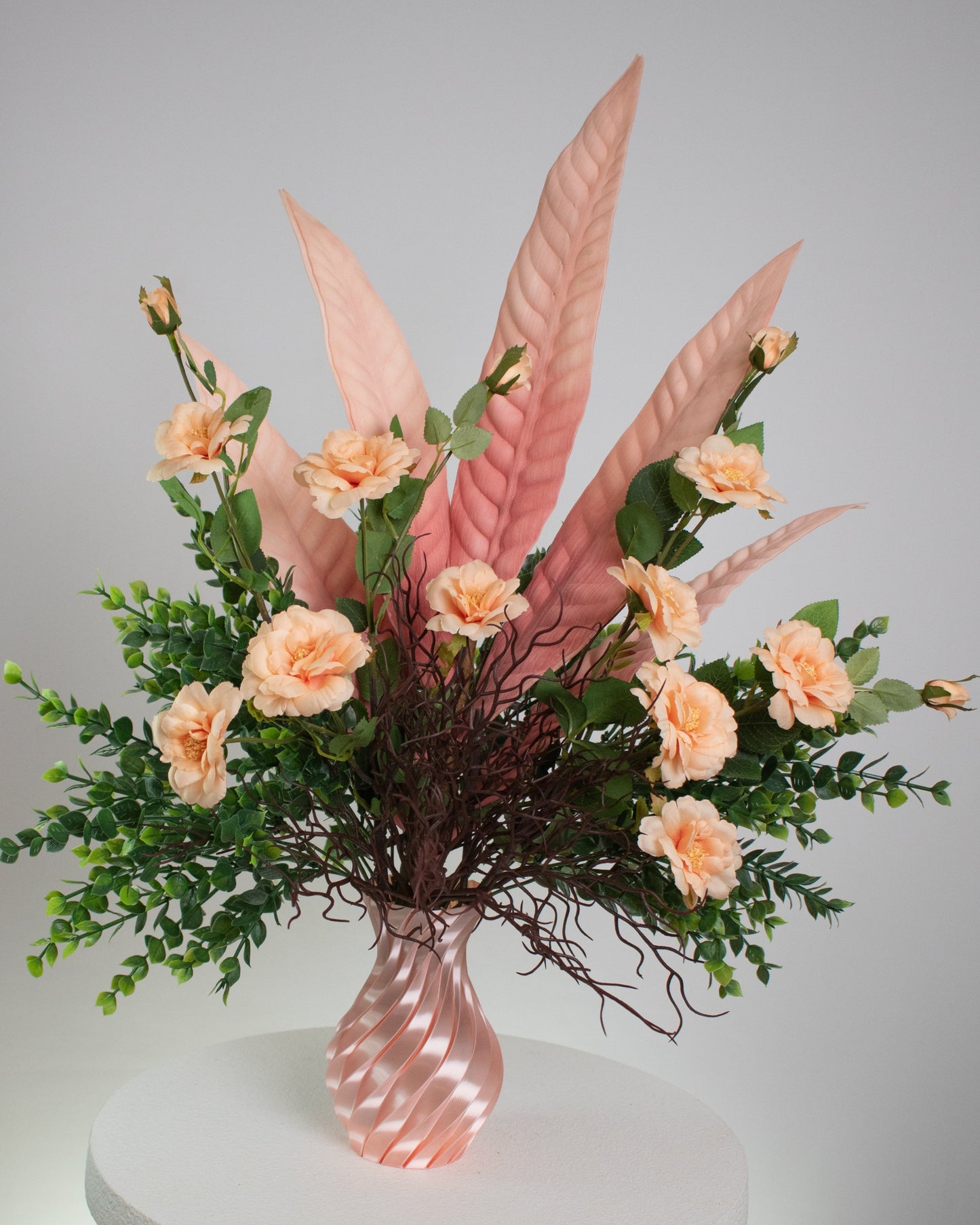 Artificial floral arrangement with peach roses, pink tropical leaves, and lush greenery in a twisted pink spiral vase, displayed on a white pedestal — perfect for elegant or modern interior spaces.