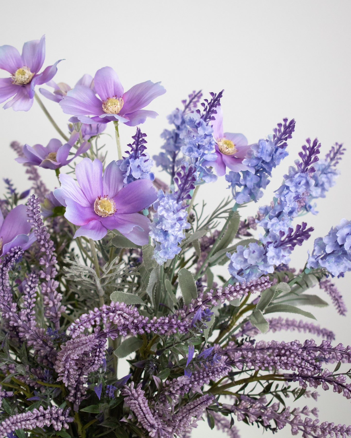Artificial lavender and lilac flower arrangement in a wavy-textured purple vase, styled on a minimalist white pedestal — a soft and elegant decorative display ideal for serene and modern interiors.