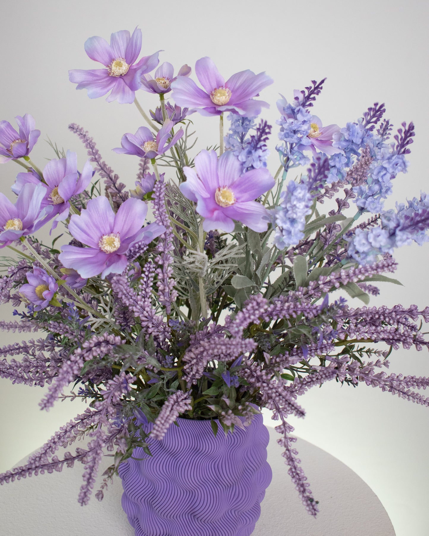 Artificial lavender and lilac flower arrangement in a wavy-textured purple vase, styled on a minimalist white pedestal — a soft and elegant decorative display ideal for serene and modern interiors.