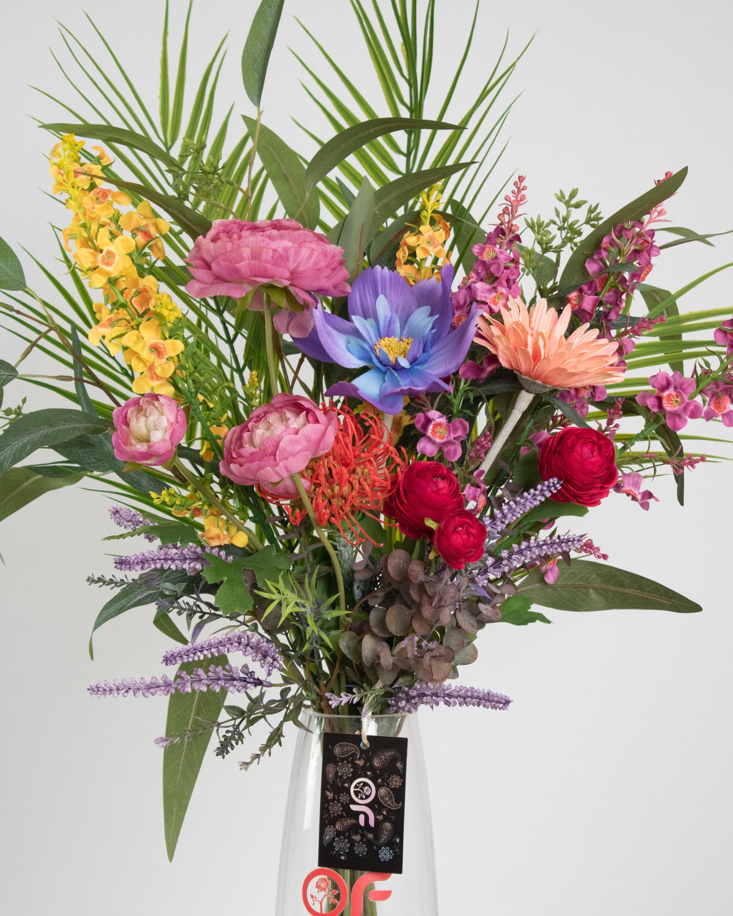 Artificial tropical floral arrangement with colourful pink, yellow, purple, orange, and blue flowers in a clear glass vase with acrylic water