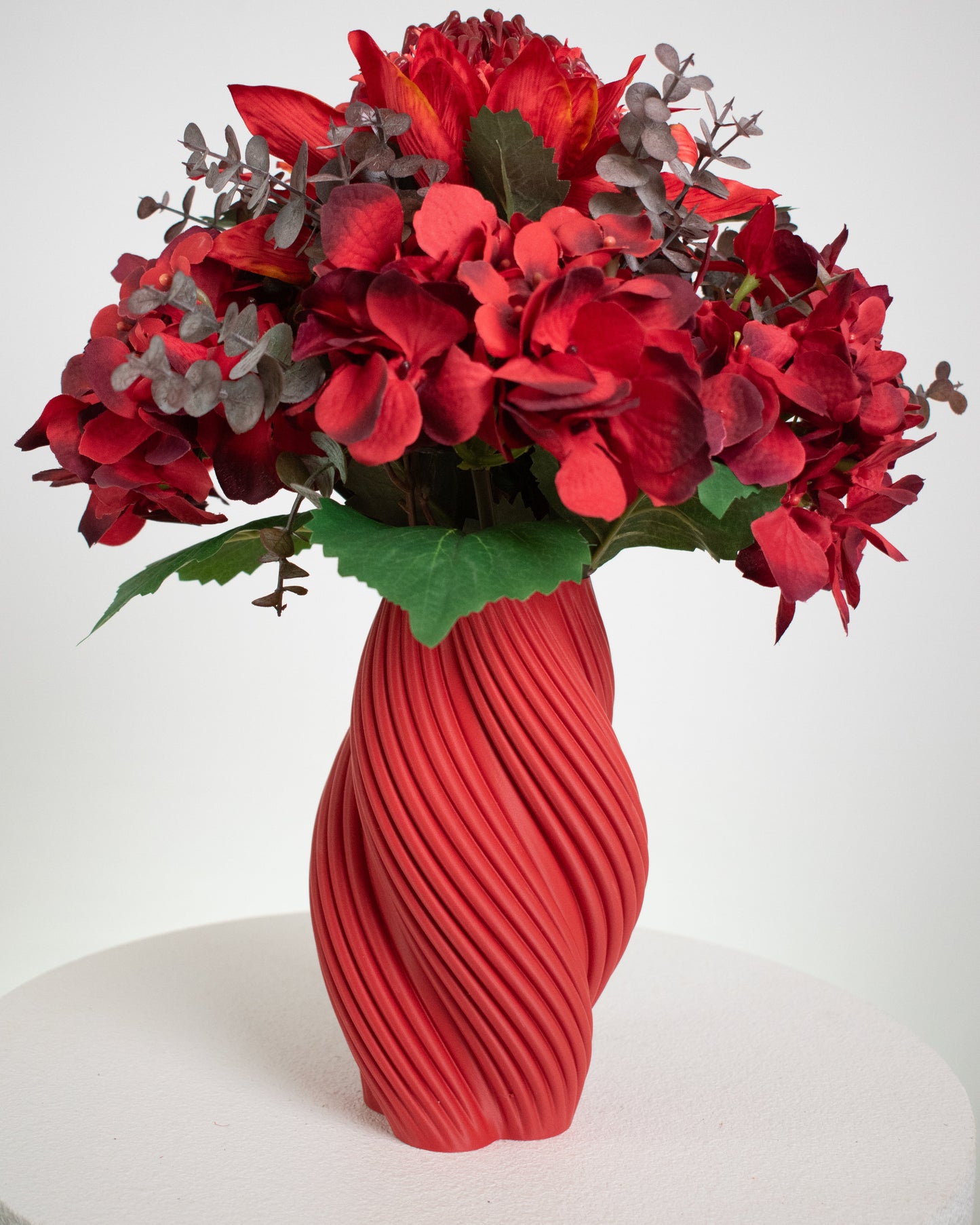 Artificial floral arrangement featuring rich red hydrangeas and crimson blooms with textured foliage, beautifully displayed in a sculptural twisted red vase.