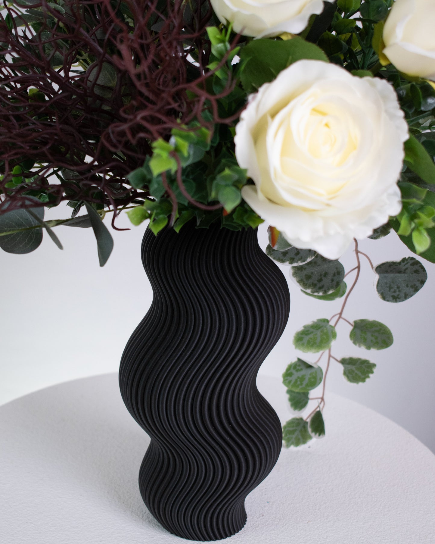 Artificial white rose arrangement with lush greenery in a sculptural matte black wavy vase, styled on a marble table — a modern and elegant decorative floral display ideal for minimalist interiors.