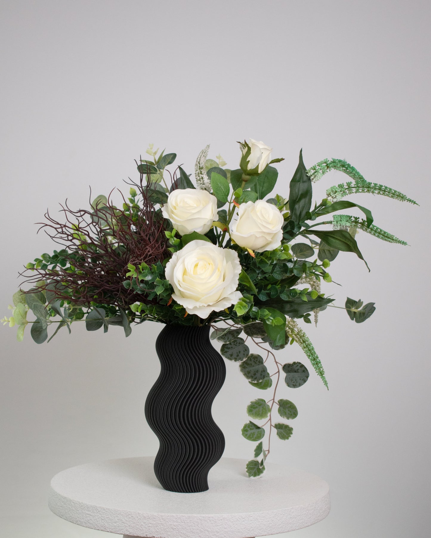 Artificial white rose arrangement with lush greenery in a sculptural matte black wavy vase, styled on a marble table — a modern and elegant decorative floral display ideal for minimalist interiors.