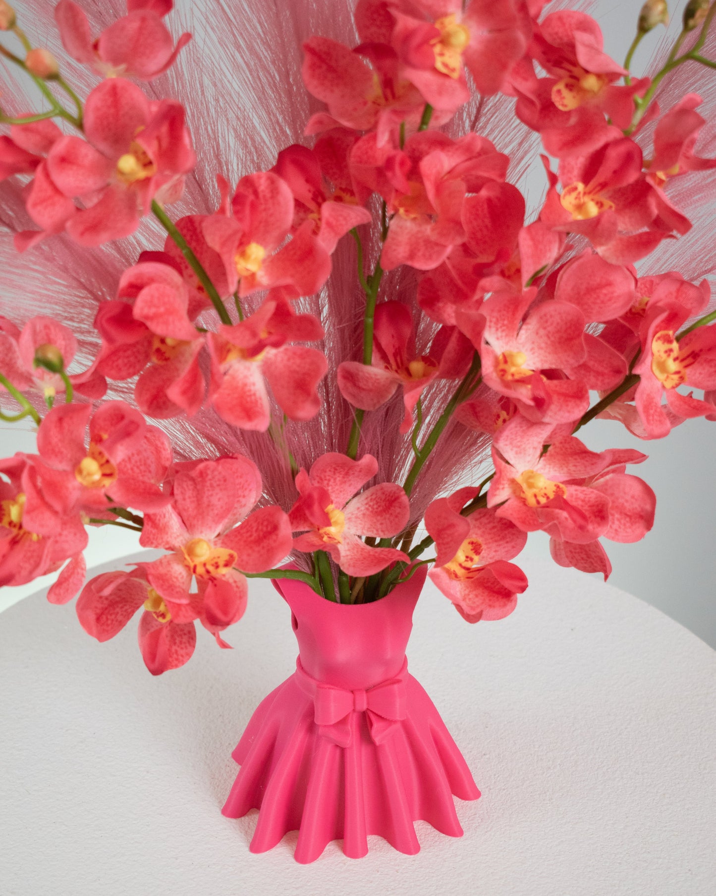 Artificial flower arrangement featuring bright pink orchids and soft pink pampas grass, beautifully styled in a sculptural pink vase shaped like an elegant dress, placed on a round white table against a light background.