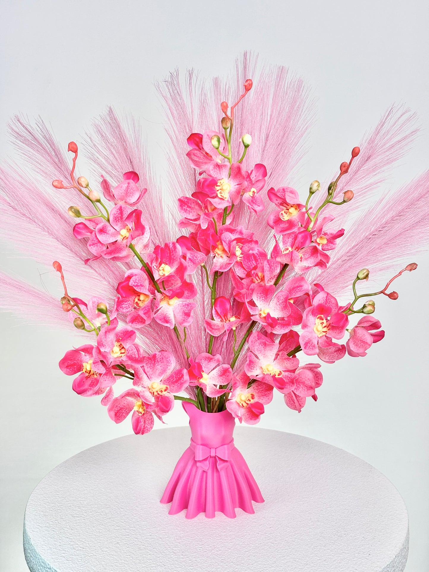 Artificial flower arrangement featuring bright pink orchids and soft pink pampas grass, beautifully styled in a sculptural pink vase shaped like an elegant dress, placed on a round white table against pink room background.
