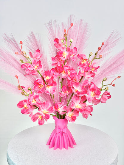 Artificial flower arrangement featuring bright pink orchids and soft pink pampas grass, beautifully styled in a sculptural pink vase shaped like an elegant dress, placed on a round white table against pink room background.