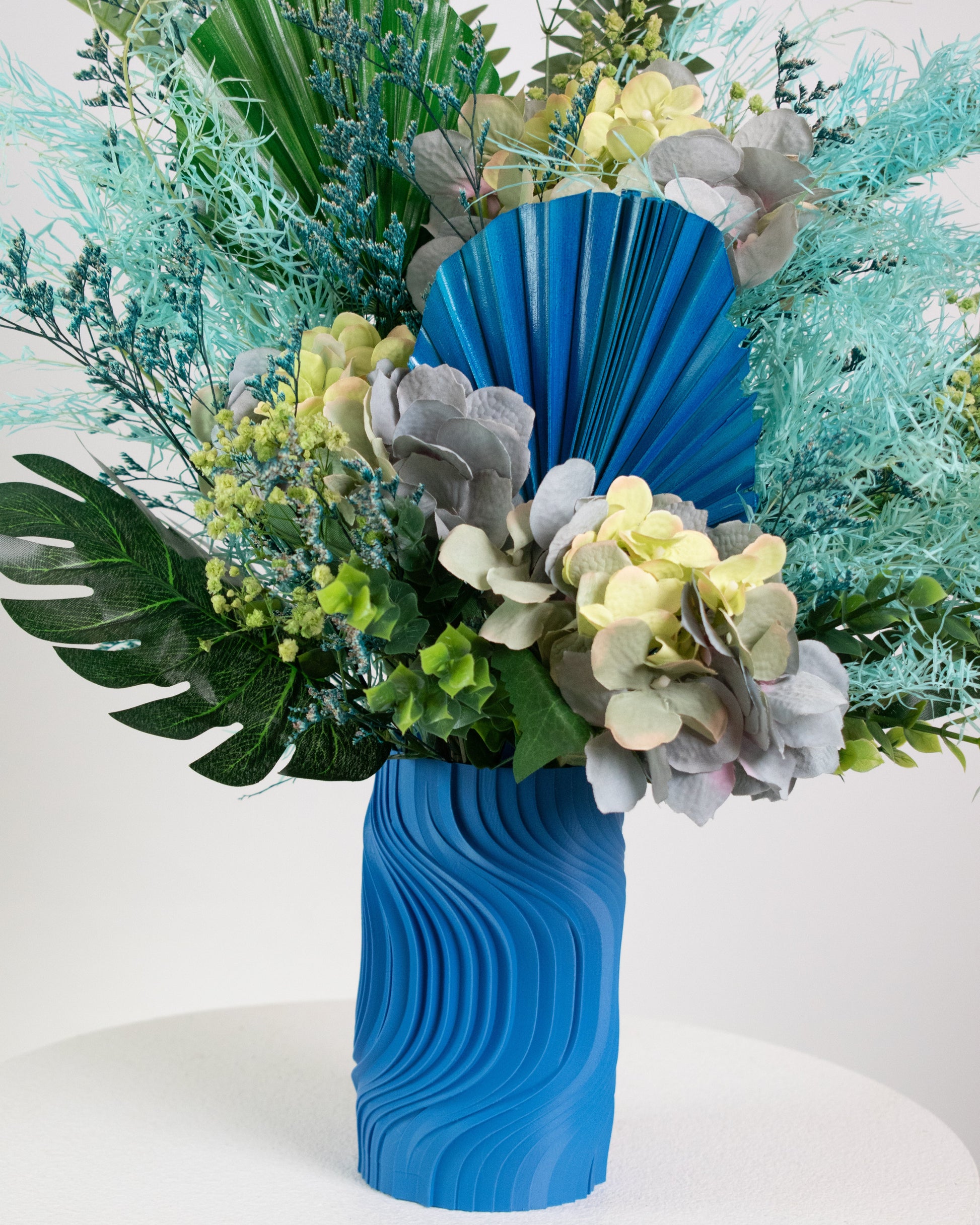 Artificial blue and green floral arrangement with tropical leaves and preserved hydrangeas in a sculptural blue vase, styled on a white pedestal against a neutral background.
