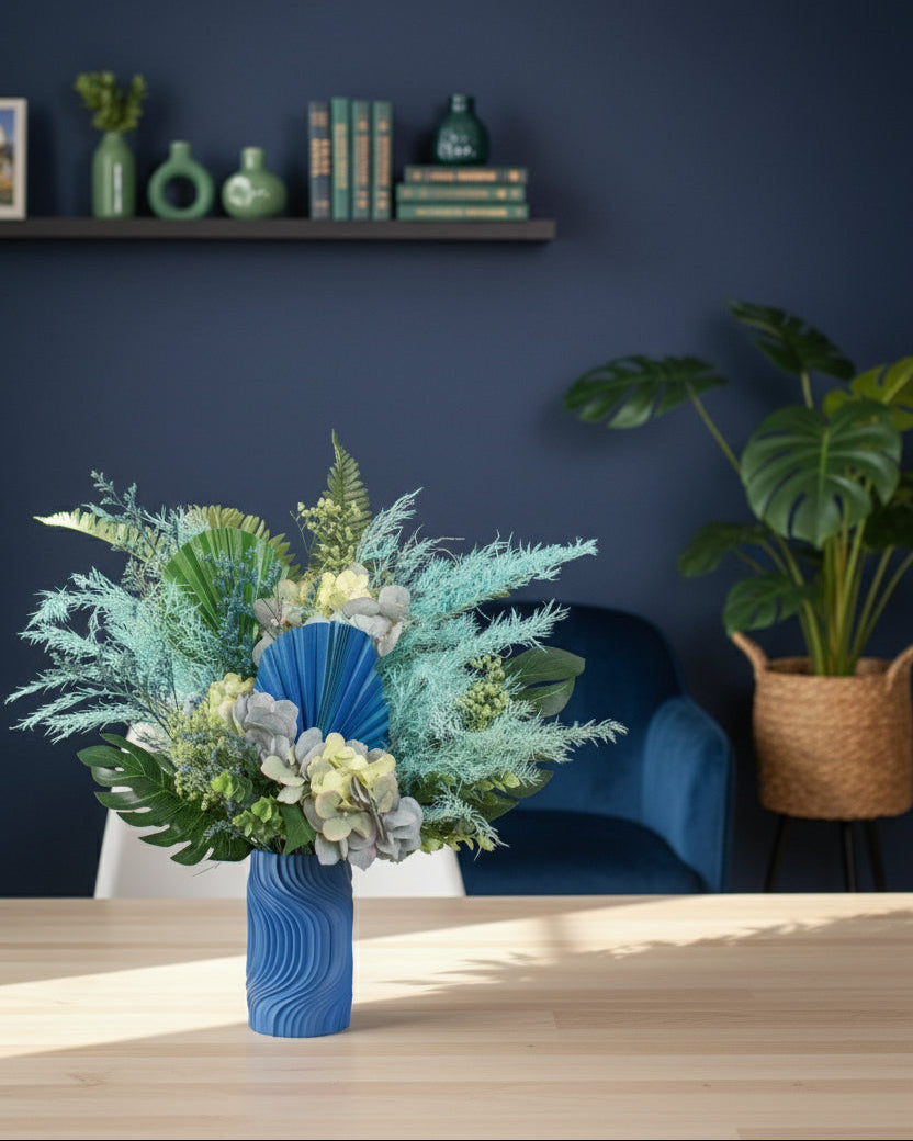 Artificial blue and green floral arrangement with tropical leaves and preserved hydrangeas in a sculptural blue vase, styled on a white pedestal against a neutral background.