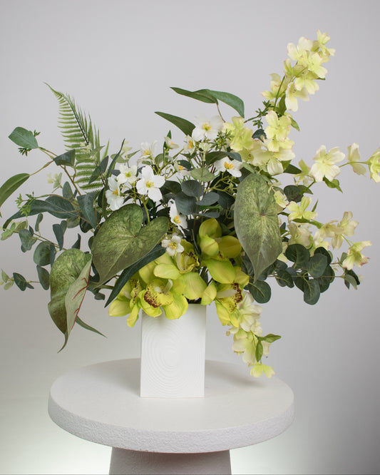 Artificial flower arrangement featuring yellow orchids, white blossoms, and lush green foliage with ferns and eucalyptus leaves, displayed in a modern white cube vase on a round white table against a neutral background.