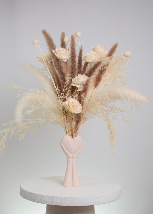 Dried floral arrangement with pampas grass and preserved flowers in neutral tones, displayed in a heart-in-hand vase