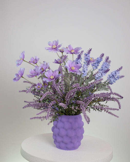 Artificial lavender and lilac flower arrangement in a wavy-textured purple vase, styled on a minimalist white pedestal — a soft and elegant decorative display ideal for serene and modern interiors.