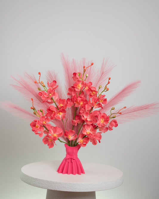 Artificial flower arrangement featuring bright pink orchids and soft pink pampas grass, beautifully styled in a sculptural pink vase shaped like an elegant dress, placed on a round white table against a light background.
