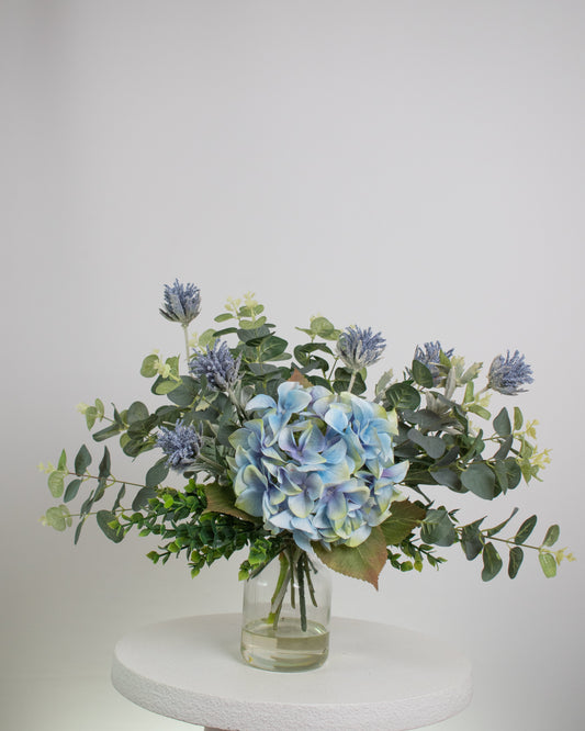 Artificial floral arrangement featuring a blue hydrangea surrounded by eucalyptus leaves and lavender-toned thistles, arranged in a clear glass vase with acrylic water, set against a neutral background.