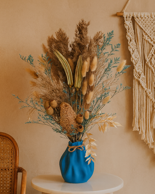 Dried floral arrangement featuring pampas grass, bunny tails, and rustic seed pods in a blue sculptural vase with jute twine, displayed on a marble pedestal.