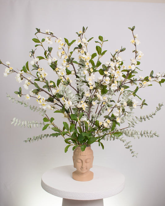 Elegant artificial floral arrangement with white blossoms and green leaves arranged in a beige Buddha head vase, symbolizing calmness and balance in modern décor.