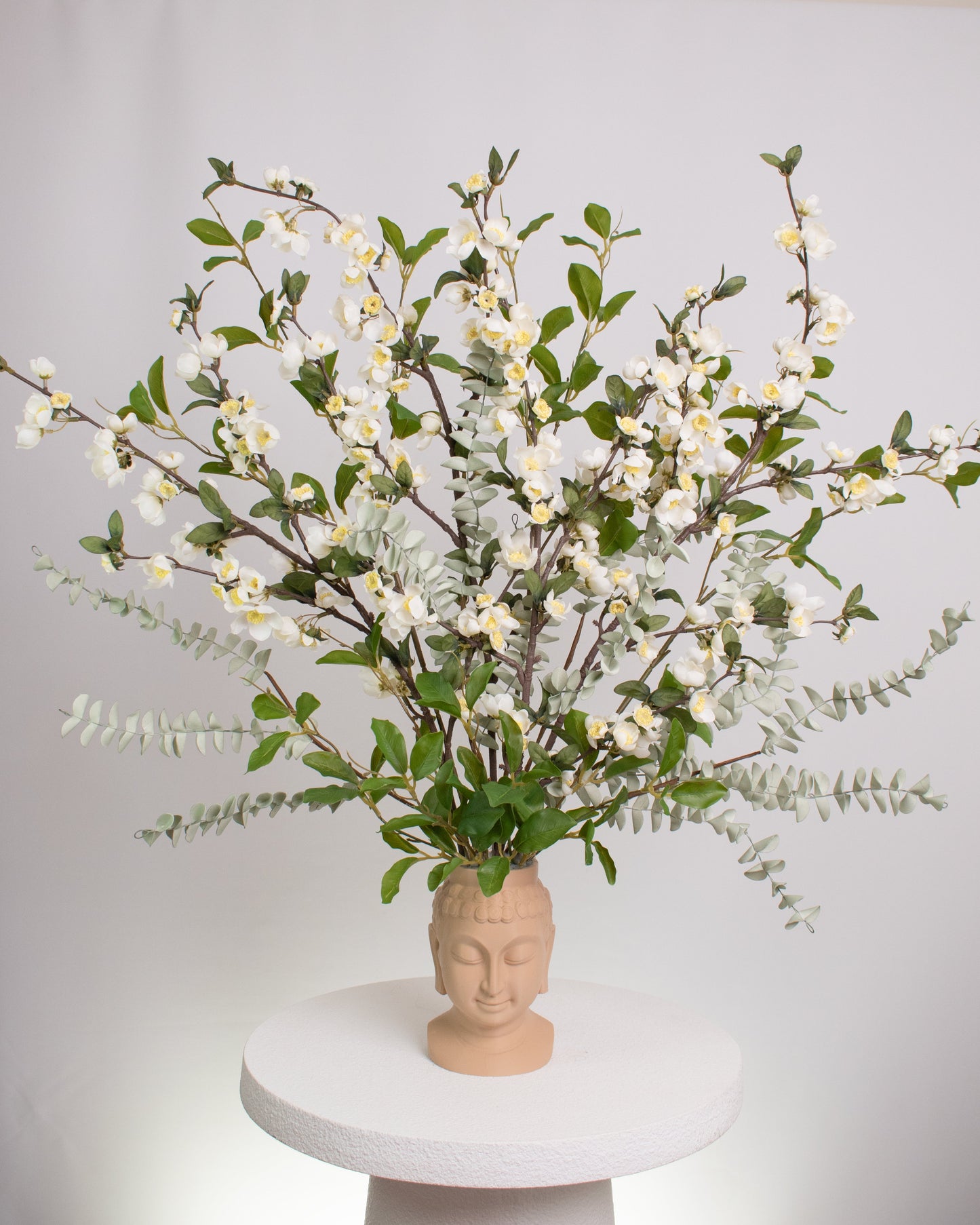 Elegant artificial floral arrangement with white blossoms and green foliage displayed in a beige Buddha head vase, symbolizing calm and mindfulness in a serene design.