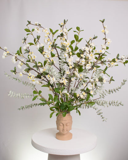 Elegant artificial floral arrangement with white blossoms and green foliage displayed in a beige Buddha head vase, symbolizing calm and mindfulness in a serene design.
