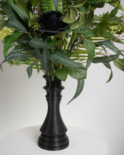 Artificial arrangement with black roses and lush green foliage displayed in a chess-queen–shaped matte-black vase, symbolizing power, sophistication, and modern elegance.