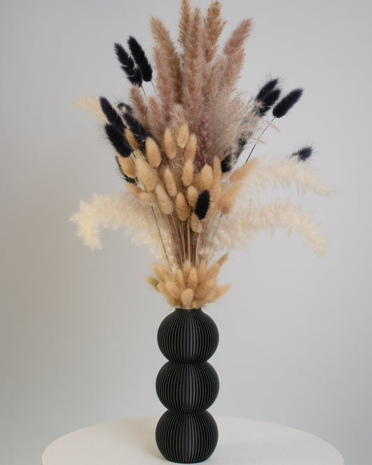 A tall arrangement of dried pampas grass and bunny tails in beige and black tones, displayed in a matte black three-sphere vase on a white table.