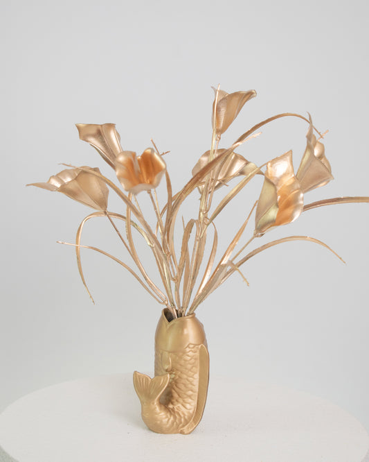 Golden metallic artificial calla lilies arranged in a gold fish-shaped vase, displayed on a white pedestal against a light neutral background.
