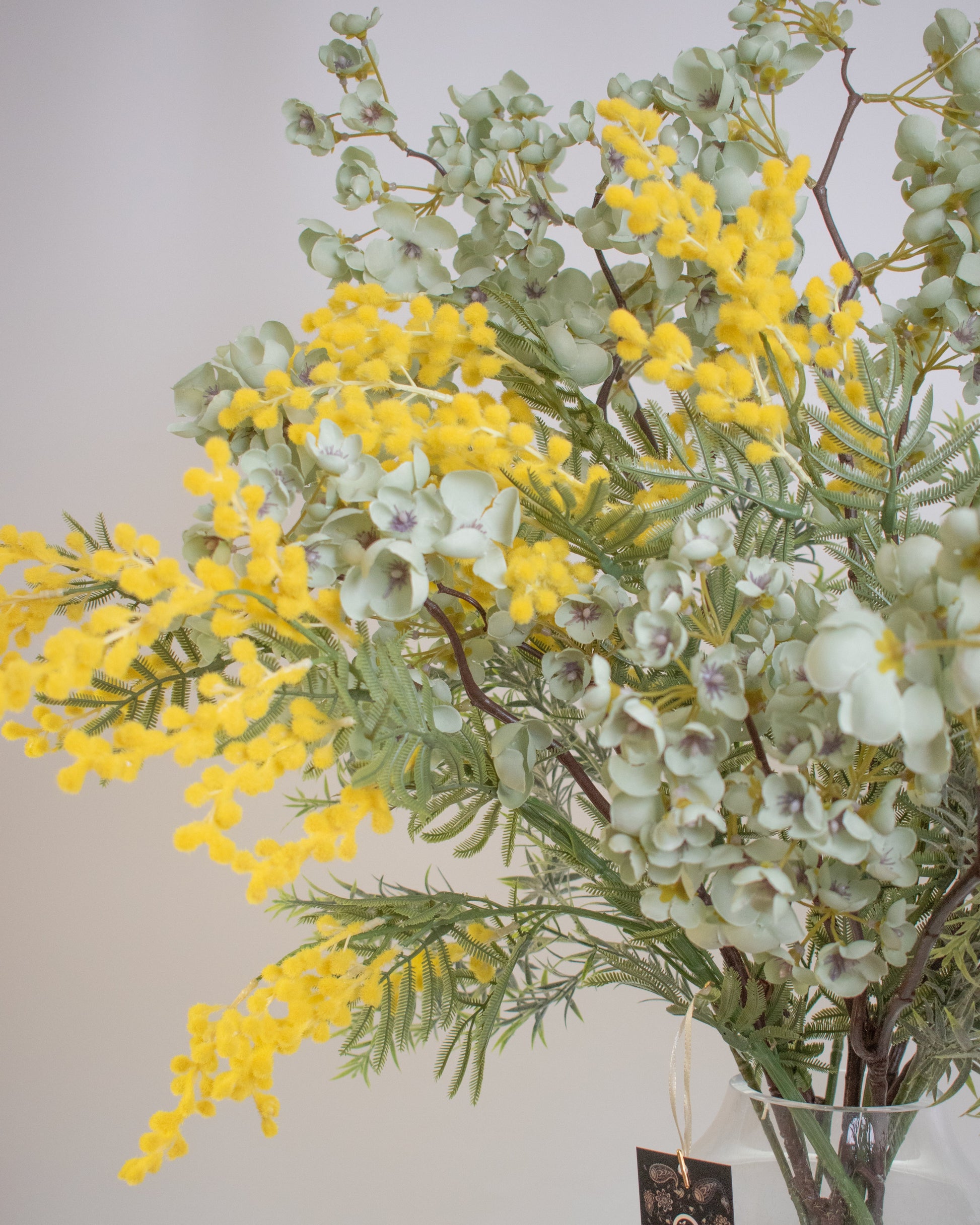 Artificial flower arrangement featuring vibrant yellow wattle blooms mixed with soft green eucalyptus leaves, displayed in a clear glass vase filled with faux stems for a natural, elegant look.
