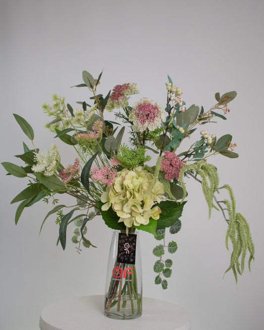 Artificial floral arrangement featuring green hydrangeas, pink wildflowers, and lush foliage in a clear glass vase with acrylic water