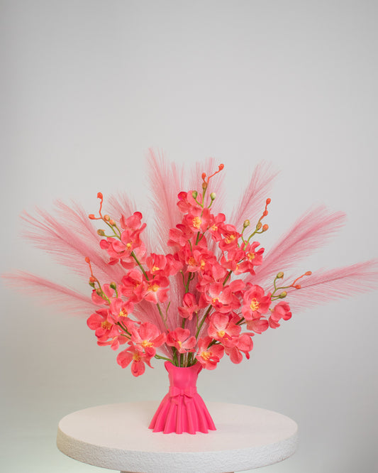 Artificial flower arrangement featuring bright pink orchids and soft pink pampas grass, beautifully styled in a sculptural pink vase shaped like an elegant dress, placed on a round white table against a light background.